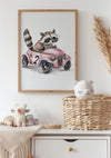 Een vintage ontwerp van een ingelijste illustratie met een wasbeer die een roze auto bestuurt met nummer 2 hangt aan een witte muur. Daaronder, in een rieten mand met een pluchen berenkop en knuffels op een wit dressoir, is op de achtergrond pampagras geplaatst. Het Wasbeer Met Roze Vintage Auto Schilderij van CollageDepot maakt gebruik van een magnetisch ophangsysteem.,Lichtbruin