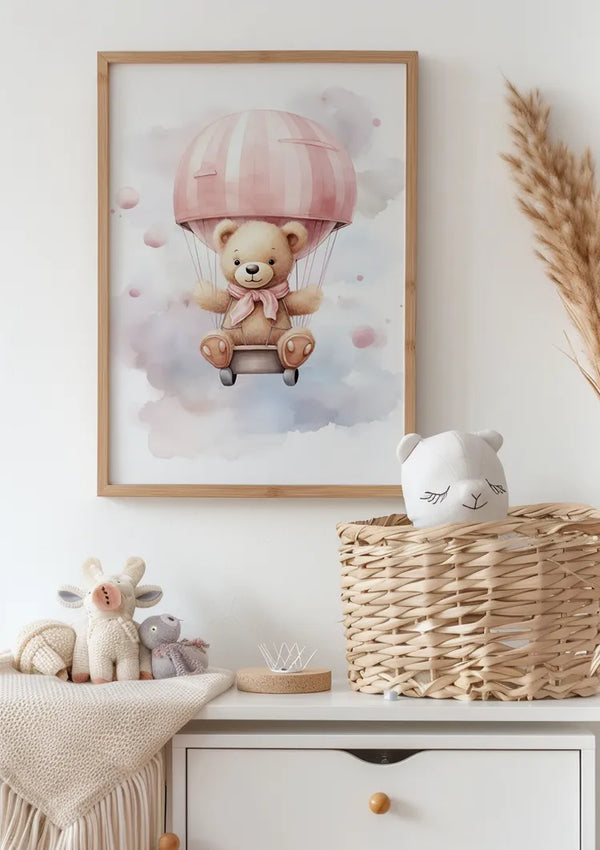 Een ingelijst **CollageDepot Teddybeer Met Roze Luchtballon Schilderij** van een beer in een ballonluchtschip hangt aan een witte muur boven een dressoir. Op het dressoir staan knuffels, een rieten mand met een opgezette berenkop en een klein dienblad. Decoratief pampagras wordt in een hoek geplaatst, wat bijdraagt aan de charmante wanddecoratie.,Lichtbruin